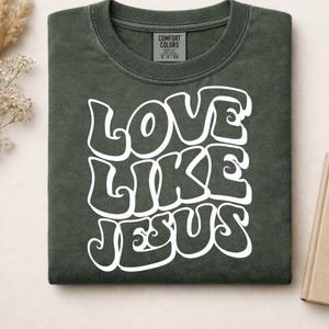 Pu&ograve; includere: Una t-shirt a girocollo verde oliva scuro con la scritta bianca LOVE LIKE JESUS in un carattere audace, ondulato e di ispirazione retr&ograve;. La maglietta presenta un'etichetta del marchio Comfort Colors all'interno del collo, indicante la taglia S.