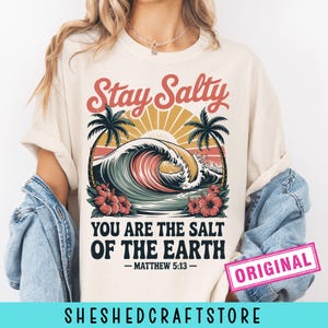 May include: A cream-coloured t-shirt with a graphic of an ocean wave, palm trees, and the text "Stay Salty." The text "You Are The Salt Of The Earth - Matthew 5:13" is below. A pink "ORIGINAL" stamp is visible on the bottom right.