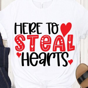 May include: White t-shirt with the phrase "HERE TO STEAL HEARTS" in black and red lettering. The word "STEAL" is in red with heart accents. A red heart graphic is above the word "HEARTS".