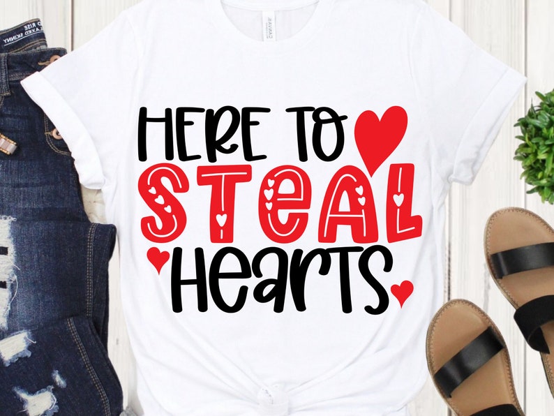 May include: White t-shirt with the phrase "HERE TO STEAL HEARTS" in black and red lettering. The word "STEAL" is in red with heart accents. A red heart graphic is above the word "HEARTS".
