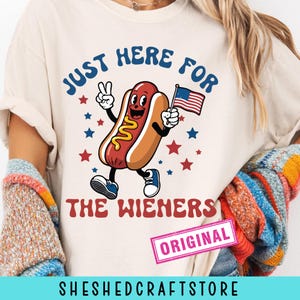 Op de afbeelding: Crèmekleurig T-shirt met een cartoon hotdog met een lachend gezicht, die een Amerikaanse vlag vasthoudt en een vredesteken geeft. De tekst luidt "JUST HERE FOR THE WIENERS" in blauw en rood. Sterren omringen de hotdog.