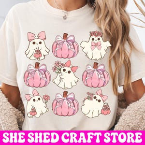 K&ouml;nnte beinhalten: Ein cremefarbenes T-Shirt mit einem Grafikdruck von drei wei&szlig;en Geistern und vier rosa K&uuml;rbissen in einem Raster. Die Geister sind mit rosa Schleifen und Blumen verziert, w&auml;hrend die K&uuml;rbisse mit gro&szlig;en, halbtransparenten rosa Schleifen geschm&uuml;ckt sind.