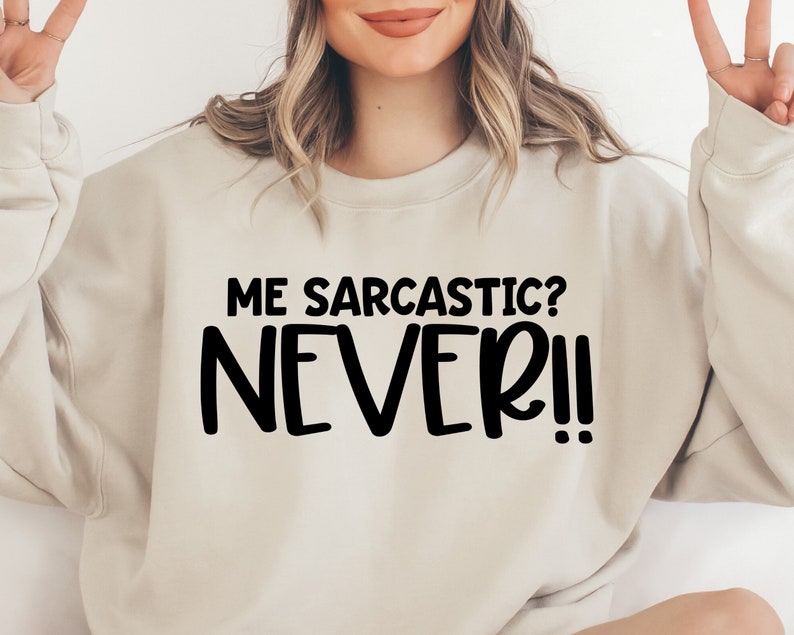 May include: A light beige sweatshirt with the text "ME SARCASTIC? NEVER!" printed in black.