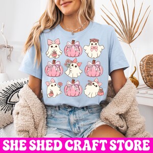 K&ouml;nnte beinhalten: Ein hellblaues T-Shirt mit einem Rastermuster aus neun Illustrationen. Das Design zeigt wei&szlig;e Geister mit rosa Schleifen und Blumen, abwechselnd mit rosa K&uuml;rbissen, die mit Schleifen verziert sind. Die Motive haben eine sanfte Pastellfarbpalette.