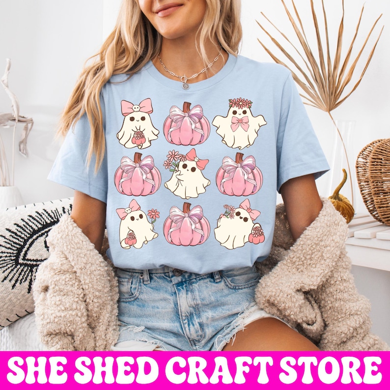 K&ouml;nnte beinhalten: Ein hellblaues T-Shirt mit einem Rastermuster aus neun Illustrationen. Das Design zeigt wei&szlig;e Geister mit rosa Schleifen und Blumen, abwechselnd mit rosa K&uuml;rbissen, die mit Schleifen verziert sind. Die Motive haben eine sanfte Pastellfarbpalette.