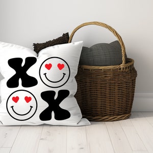 Pu&ograve; includere: Cuscino bianco con XO nero e due facce sorridenti con cuori rossi per gli occhi. Il cuscino &egrave; su un pavimento di legno bianco accanto a un cesto di vimini con una fodera in tessuto grigio.
