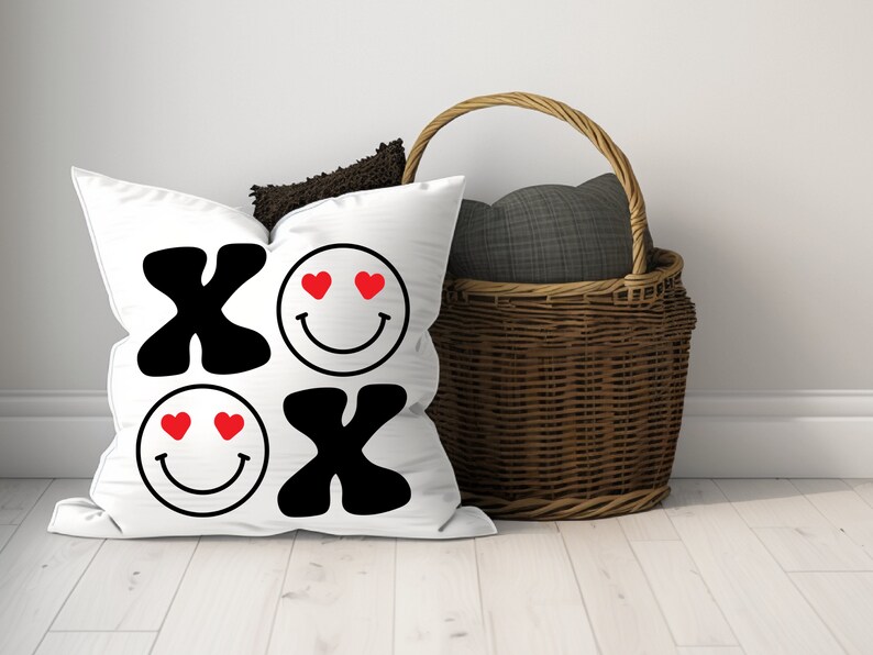 Pu&ograve; includere: Cuscino bianco con XO nero e due facce sorridenti con cuori rossi per gli occhi. Il cuscino &egrave; su un pavimento di legno bianco accanto a un cesto di vimini con una fodera in tessuto grigio.