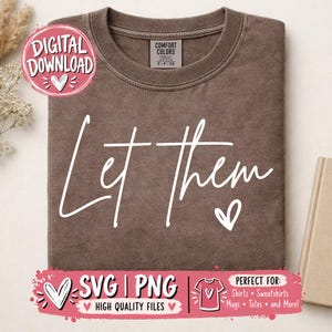 Può includere: Una t-shirt marrone a girocollo con la scritta bianca Let Them in carattere corsivo, accompagnata da un piccolo cuore bianco. La maglietta presenta un'etichetta del marchio Comfort Colors all'interno del colletto e un tessuto testurizzato.