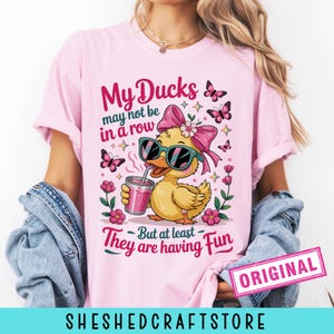 K&ouml;nnte beinhalten: Ein hellrosa T-Shirt mit einer Grafik einer gelben Ente, die eine Sonnenbrille und eine rosa Schleife tr&auml;gt und ein rosa Getr&auml;nk h&auml;lt. Der Text lautet: My Ducks may not be in a row But at least They are having Fun. Rosa Schmetterlinge und Blumen umgeben die Ente.