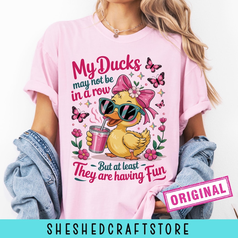 K&ouml;nnte beinhalten: Ein hellrosa T-Shirt mit einer Grafik einer gelben Ente, die eine Sonnenbrille und eine rosa Schleife tr&auml;gt und ein rosa Getr&auml;nk h&auml;lt. Der Text lautet: My Ducks may not be in a row But at least They are having Fun. Rosa Schmetterlinge und Blumen umgeben die Ente.