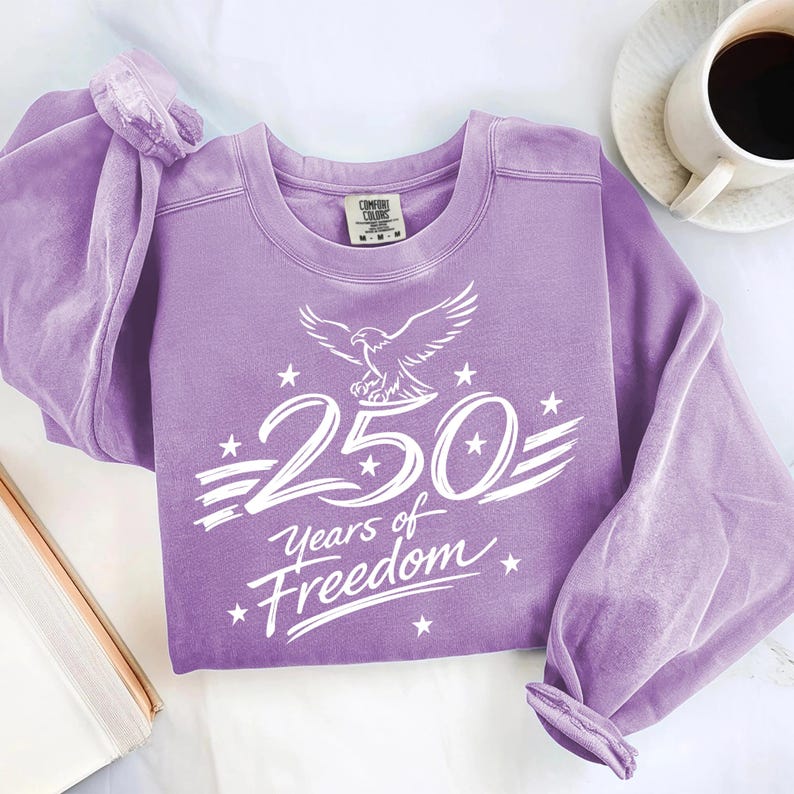 K&ouml;nnte beinhalten: Ein lang&auml;rmeliges, lavendelfarbenes Sweatshirt mit Rundhalsausschnitt und einem wei&szlig;en Grafikdruck eines Adlers &uuml;ber dem Text 250 Years of Freedom, verziert mit Sternen und Streifen. Das Kleidungsst&uuml;ck hat ein Comfort Colors Markenetikett.