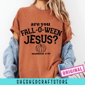 May include: A burnt orange t-shirt with the text "are you FALL-O-WEEN JESUS?" in black, along with a pumpkin graphic and "matthew 4:19". The shirt has rolled-up sleeves and is paired with a denim jacket. The word "ORIGINAL" is in a pink rectangle.