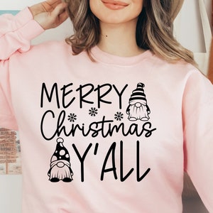May include: A pink sweatshirt with the text "Merry Christmas Y'all" in black lettering. Two gnomes are featured on the sweatshirt, one with a black and white striped hat and the other with a black and white polka dot hat.