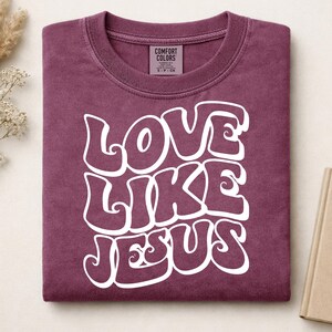 Pu&ograve; includere: Una t-shirt a maniche corte color bordeaux con scollo rotondo e un'etichetta del marchio Comfort Colors all'interno del colletto. La parte anteriore presenta la scritta bianca LOVE LIKE JESUS in un carattere grande, audace e ondulato.
