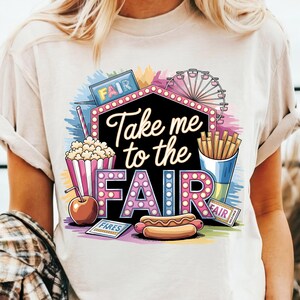 May include: White t-shirt with a colorful graphic design featuring a carnival scene. The design includes a Ferris wheel, popcorn, hot dogs, french fries, and a sign that says "Take me to the fair".