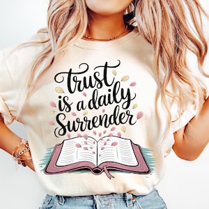 May include: Cream-colored sweatshirt with the text "Trust is a daily Surrender" in black script. The design includes an open book with pink pages and falling petals. The overall design is a soft, inspirational graphic.