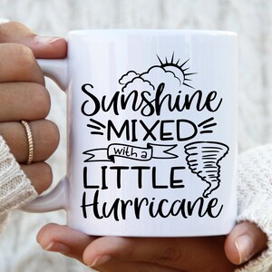May include: White ceramic mug with the text "Sunshine Mixed with a Little Hurricane" in black script. The design includes a sun, clouds, and a tornado illustration. The mug has a curved handle.