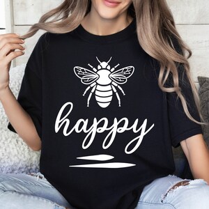 May include: Black t-shirt with a white graphic of a bee and the word "happy" in cursive script. The bee design is detailed with wings and body segments. Below the word "happy" are two horizontal lines.