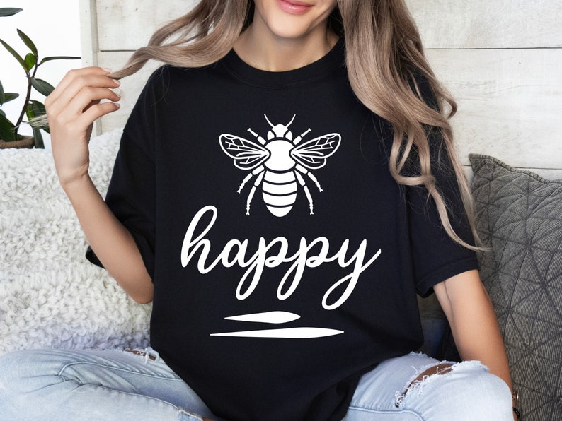 May include: Black t-shirt with a white graphic of a bee and the word "happy" in cursive script. The bee design is detailed with wings and body segments. Below the word "happy" are two horizontal lines.