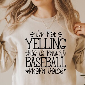 May include: A beige sweatshirt with black text that reads "I'm not yelling this is my baseball mom voice" with a black baseball graphic.