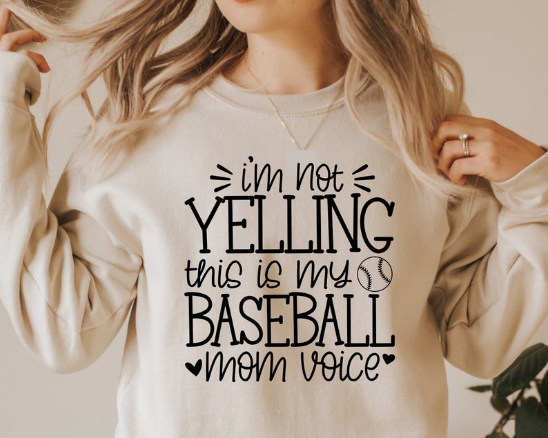 May include: A beige sweatshirt with black text that reads "I'm not yelling this is my baseball mom voice" with a black baseball graphic.