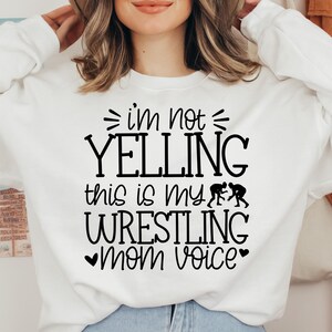 May include: White sweatshirt with black text that reads "I'm not yelling this is my wrestling mom voice" with a heart on either side of the word "mom". There are two black silhouettes of wrestlers on the shirt.