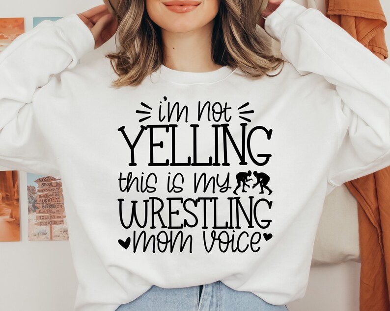 May include: White sweatshirt with black text that reads "I'm not yelling this is my wrestling mom voice" with a heart on either side of the word "mom". There are two black silhouettes of wrestlers on the shirt.