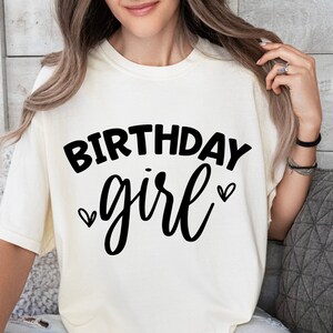 May include: A white t-shirt with the text "Birthday girl" in black lettering and two small hearts on either side of the word "girl".