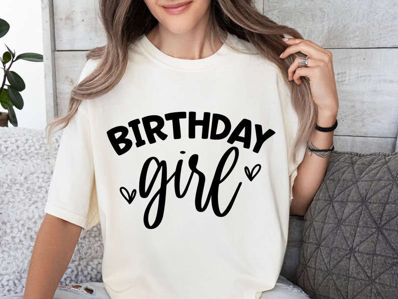 May include: A white t-shirt with the text "Birthday girl" in black lettering and two small hearts on either side of the word "girl".