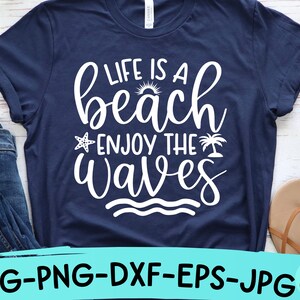 以下が含まれることがあります： 白い文字で「Life is a beach enjoy the waves」と書かれたネイビーブルーのTシャツ。テキストは筆記体で、小さな太陽、ヒトデ、ヤシの木のグラフィックが含まれています。