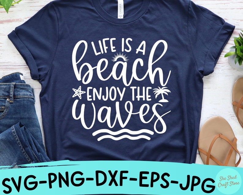 以下が含まれることがあります： 白い文字で「Life is a beach enjoy the waves」と書かれたネイビーブルーのTシャツ。テキストは筆記体で、小さな太陽、ヒトデ、ヤシの木のグラフィックが含まれています。