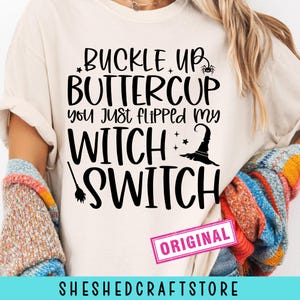 May include: A cream-colored t-shirt with the phrase "BUCKLE UP BUTTERCUP you just FLIPPED my WITCH SWITCH" in black lettering. The design includes a witch hat, stars, and a spider. The word "ORIGINAL" is in a pink rectangle.