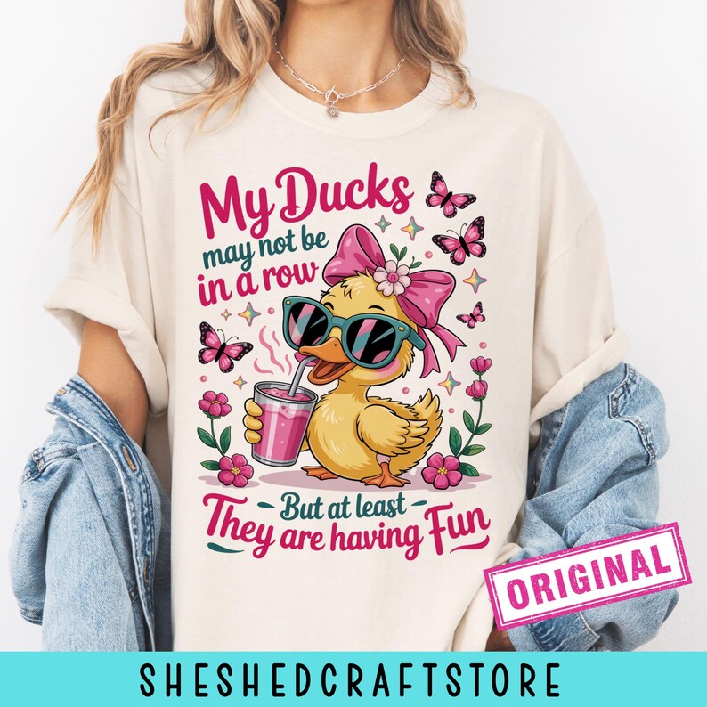 K&ouml;nnte beinhalten: Ein cremefarbenes T-Shirt mit einer Grafik einer gelben Ente, die eine Sonnenbrille und eine rosa Schleife tr&auml;gt und ein rosa Getr&auml;nk h&auml;lt. Der Text lautet: My Ducks may not be in a row But at least They are having Fun. Rosa Blumen und Schmetterlinge umgeben die Ente.