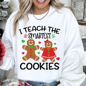 May include: White sweatshirt featuring the text "I TEACH THE SMARTEST COOKIES" in black, with gingerbread people and festive accents. A holiday-themed garment, perfect for the festive season.