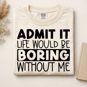 May include: A cream-colored crew neck t-shirt with black text that reads ADMIT IT LIFE WOULD BE BORING WITHOUT ME. The shirt features a Comfort Colors brand label at the inner neckline and is folded neatly on a flat surface.