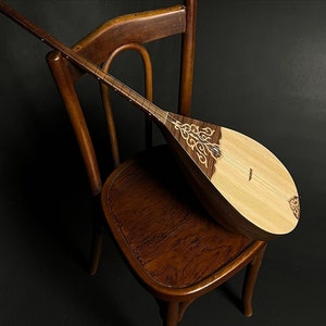 Può includere: Uno strumento musicale in legno, probabilmente un liuto, è appoggiato su una sedia vintage in legno. Lo strumento presenta una tavola armonica di colore chiaro e un corpo in legno scuro e ornato con intarsi decorativi. La sedia ha uno schienale curvo e una finitura marrone caldo.