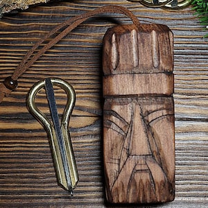 May include: A carved wooden pendant with a face design and a brass jaw harp. The pendant is brown with a leather cord. The jaw harp is gold and silver. The items are on a wooden surface.