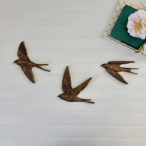 May include: Three bronze-colored swallow wall art pieces are arranged on a white, horizontally-paneled surface. The birds are in flight, with outstretched wings. A book and a flower arrangement are in the upper right corner.