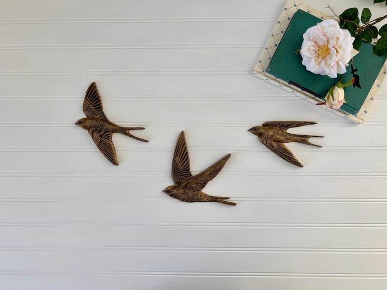 May include: Three bronze-colored swallow wall art pieces are arranged on a white, horizontally-paneled surface. The birds are in flight, with outstretched wings. A book and a flower arrangement are in the upper right corner.
