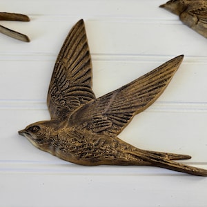 May include: Bronze-colored bird wall art in flight, with detailed wing and feather textures. The bird is mounted on a white, paneled surface. Decorative home decor.