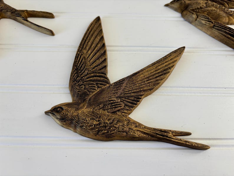 May include: Bronze-colored bird wall art in flight, with detailed wing and feather textures. The bird is mounted on a white, paneled surface. Decorative home decor.