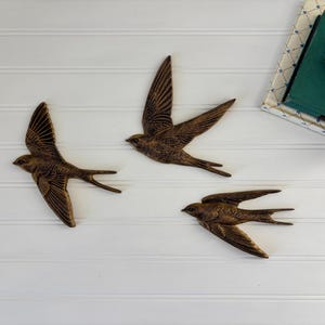 May include: Three bronze-colored swallow wall art pieces in flight, mounted on a white, horizontal-paneled surface. The birds have outstretched wings and detailed feathering. A stack of books and a flower are in the upper right corner.