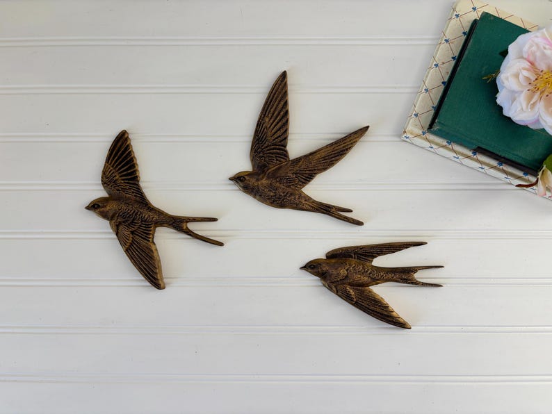 May include: Three bronze-colored swallow wall art pieces in flight, mounted on a white, horizontal-paneled surface. The birds have outstretched wings and detailed feathering. A stack of books and a flower are in the upper right corner.