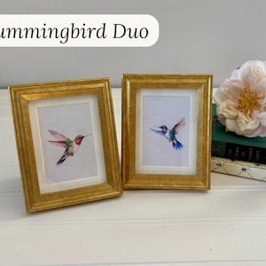 May include: Two framed hummingbird art prints. The frames are gold-colored with a rectangular shape. The art prints feature watercolor paintings of hummingbirds in flight, showcasing vibrant colors of red, green, blue, and pink. The text "Hummingbird Duo" is at the top.