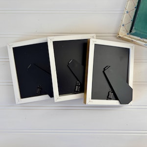 May include: Three rectangular picture frames with white borders and black backing are arranged on a white surface. The frames have black easel backs for tabletop display. A decorative book and a white flower are in the background.