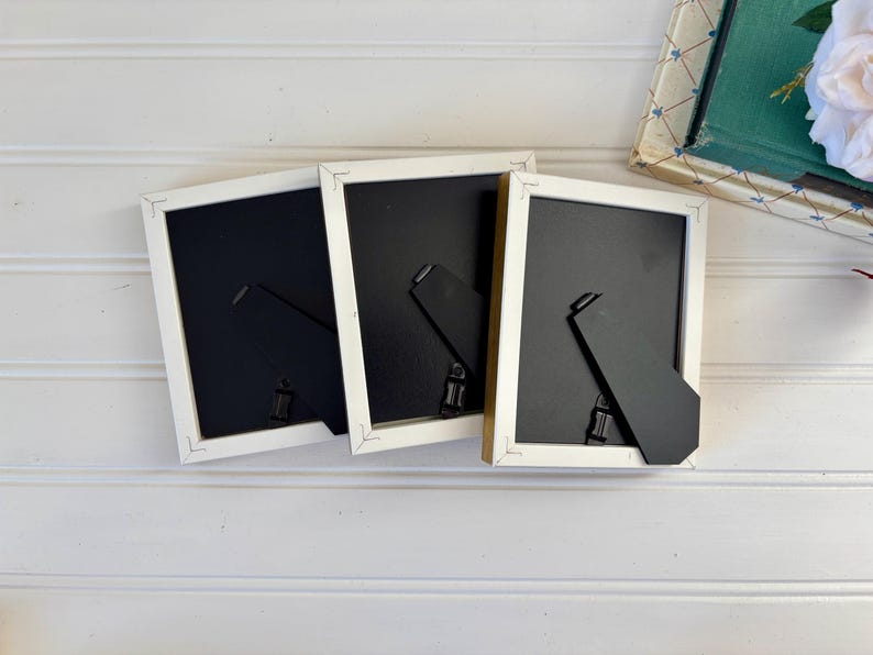 May include: Three rectangular picture frames with white borders and black backing are arranged on a white surface. The frames have black easel backs for tabletop display. A decorative book and a white flower are in the background.