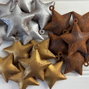 May include: A collection of metal star-shaped ornaments featuring a loop at the top for hanging. The stars are finished in three distinct textures and colors: a metallic silver, a weathered rusted brown, and a matte gold with a textured surface.