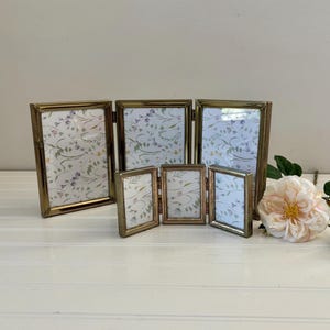 May include: A collection of antique gold-toned picture frames, including two tri-fold frames and two smaller double frames. The frames display floral patterned inserts and are arranged on a white surface, with a rose and greenery in the background.