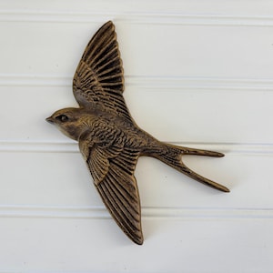 May include: A bronze-colored decorative bird in flight, with detailed wing and feather textures. The bird is mounted on a white, horizontal-paneled surface. The bird's wings are spread wide, and its tail feathers are extended.