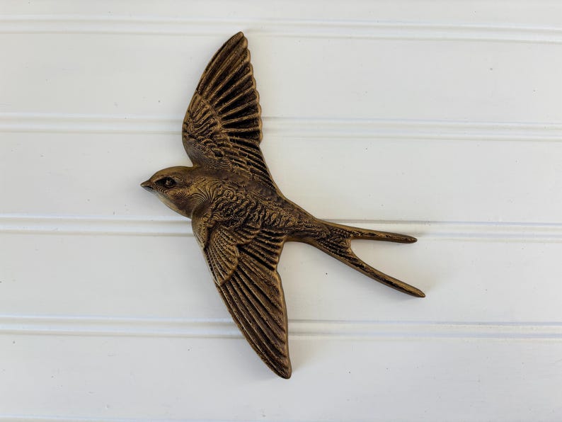 May include: A bronze-colored decorative bird in flight, with detailed wing and feather textures. The bird is mounted on a white, horizontal-paneled surface. The bird's wings are spread wide, and its tail feathers are extended.